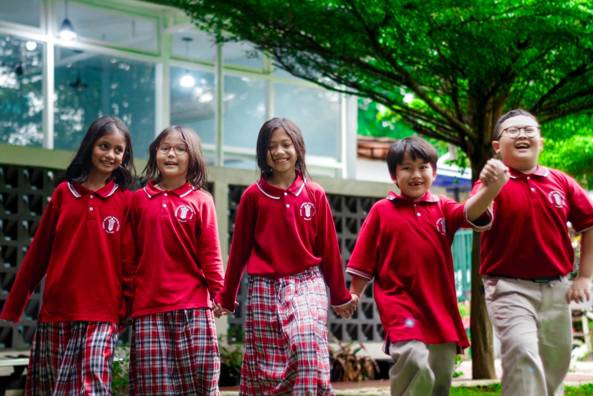 Five smiling students walking together hand in hand under a tree on campus
